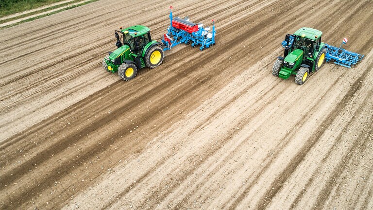 Dva traktora John Deere 6M na polju gole zemlje. Jedan vuče Monsomen sadilicu.  Drugi vuče kombinaciju Lemken gredica za sjeme