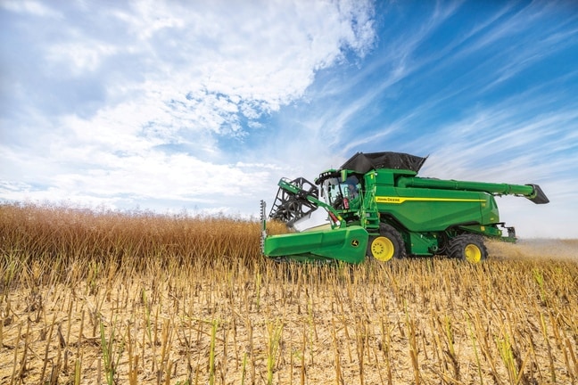 John Deere kombajn žanje zrno s hederom u polju pod plavim nebom tijekom žetve kulture