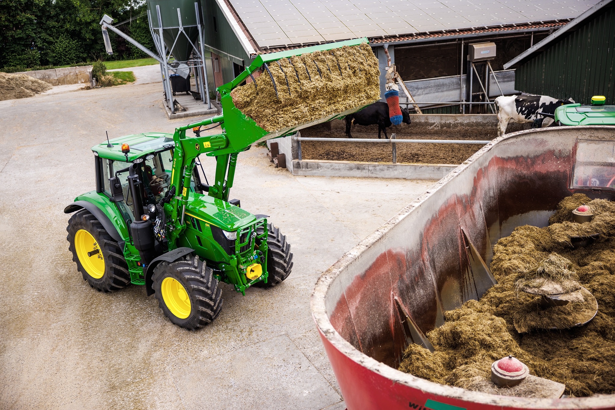 Traktor John Deere serije 6M koji prednjim utovarivačem puni silažu u miješalicu na imanju.