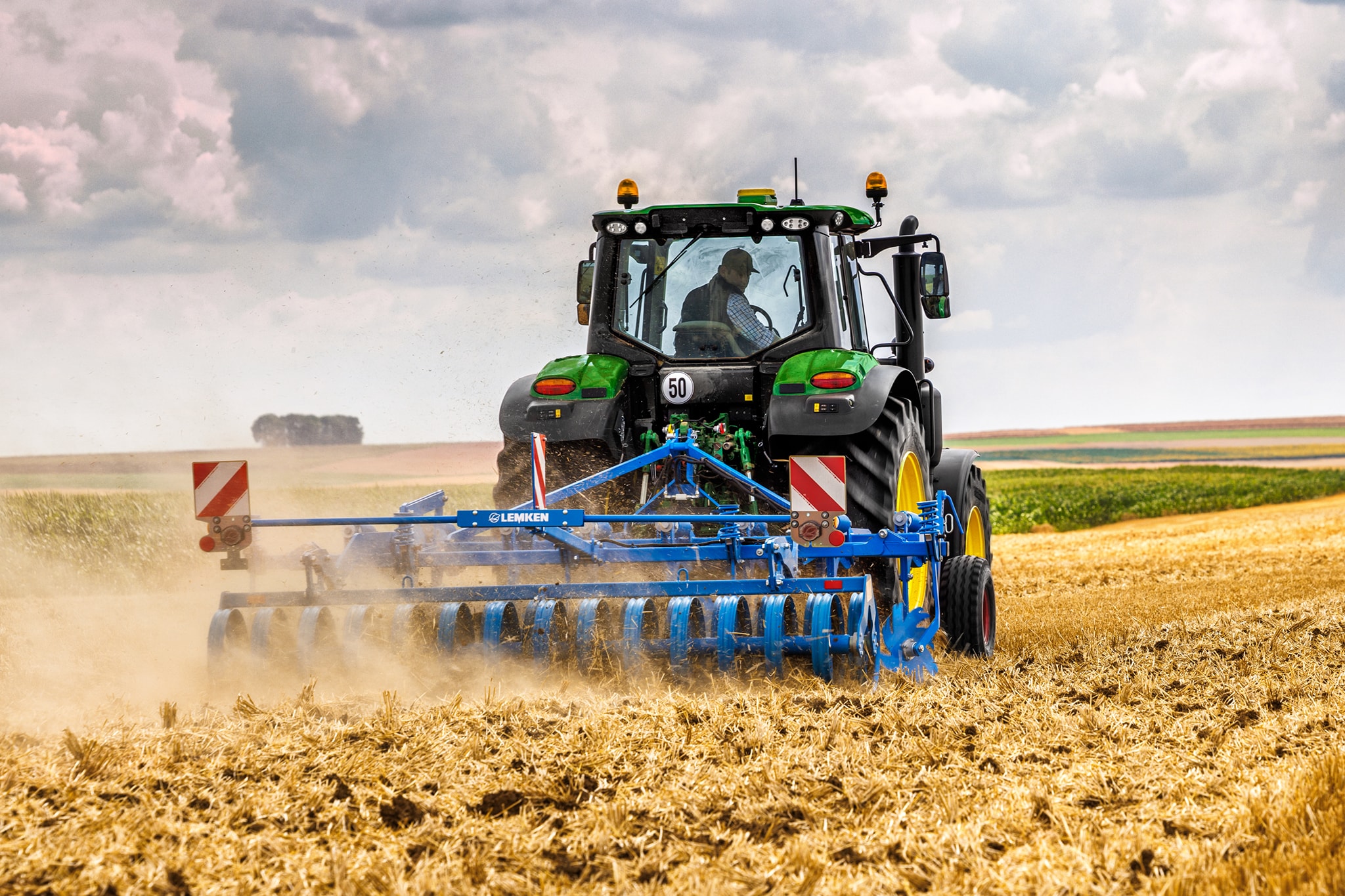 Traktor John Deere serije 6M obrađuje požnjeveno polje tanjuračom, pogled straga u prašnjavim uvjetima rada na polju.