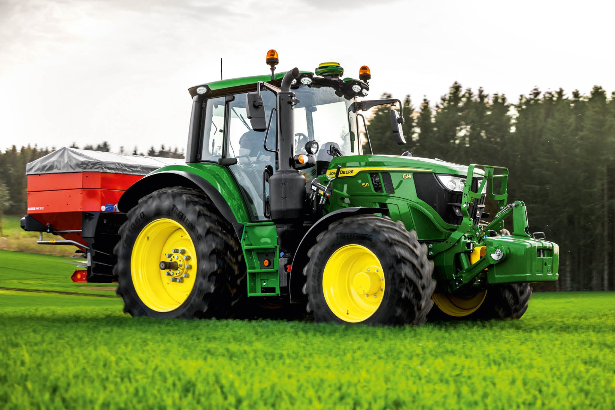 Traktor John Deere serije 6M s rasipačem montiranim straga dok radi na travi blizu ruba šume.