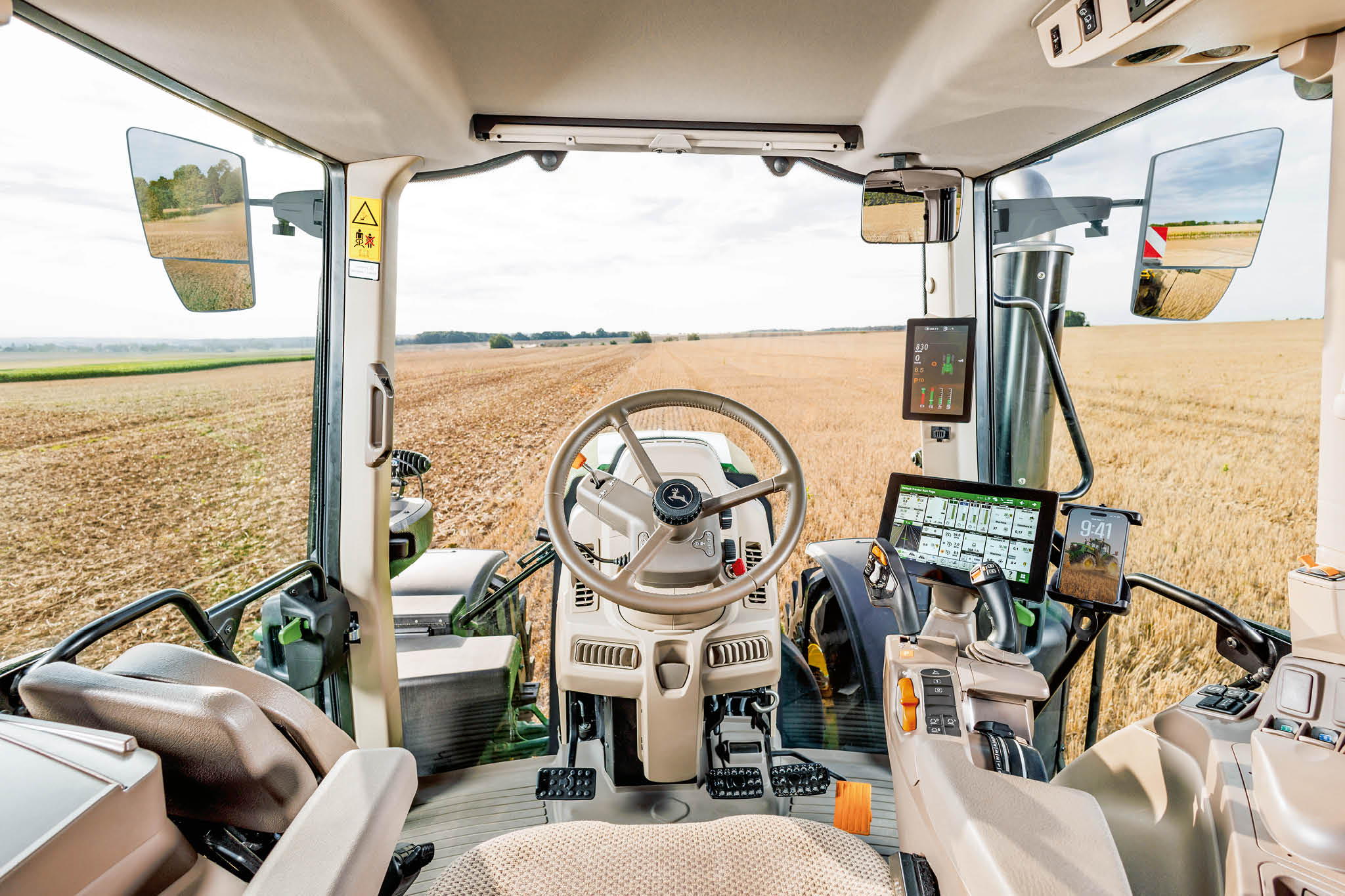 Unutrašnjost kabine traktora John Deere s kolom upravljača, digitalnim kontrolnim zaslonima i pogledom na polje kroz vjetrobransko staklo tijekom poljoprivrednih radova.