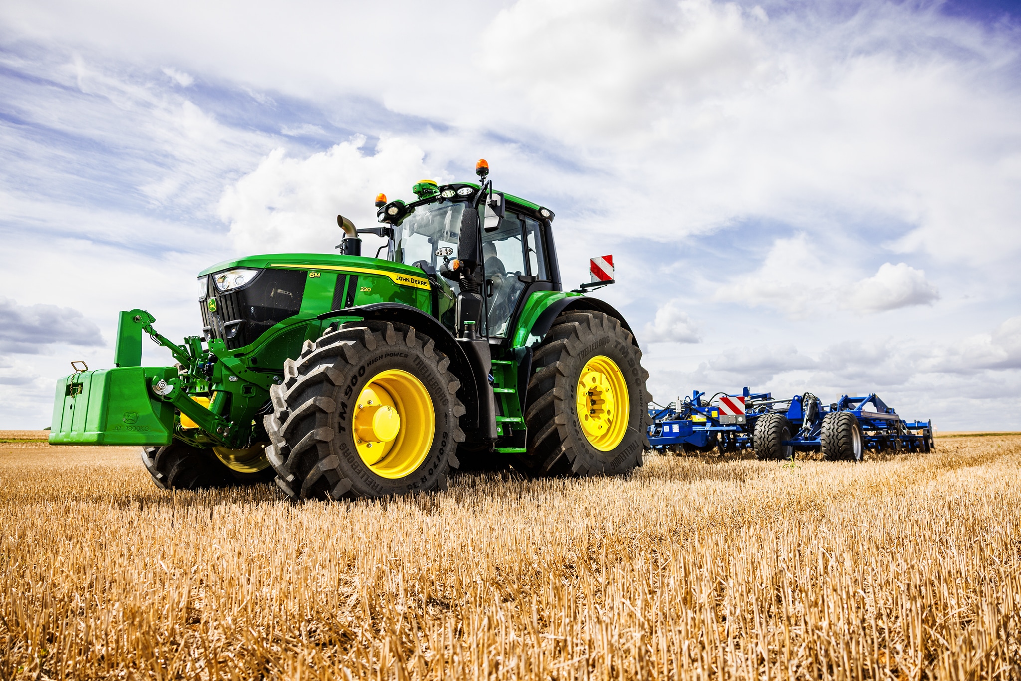 Traktor John Deere serije 6M vuče teški alat za prevrtanje po strništu pod oblačnim nebom.