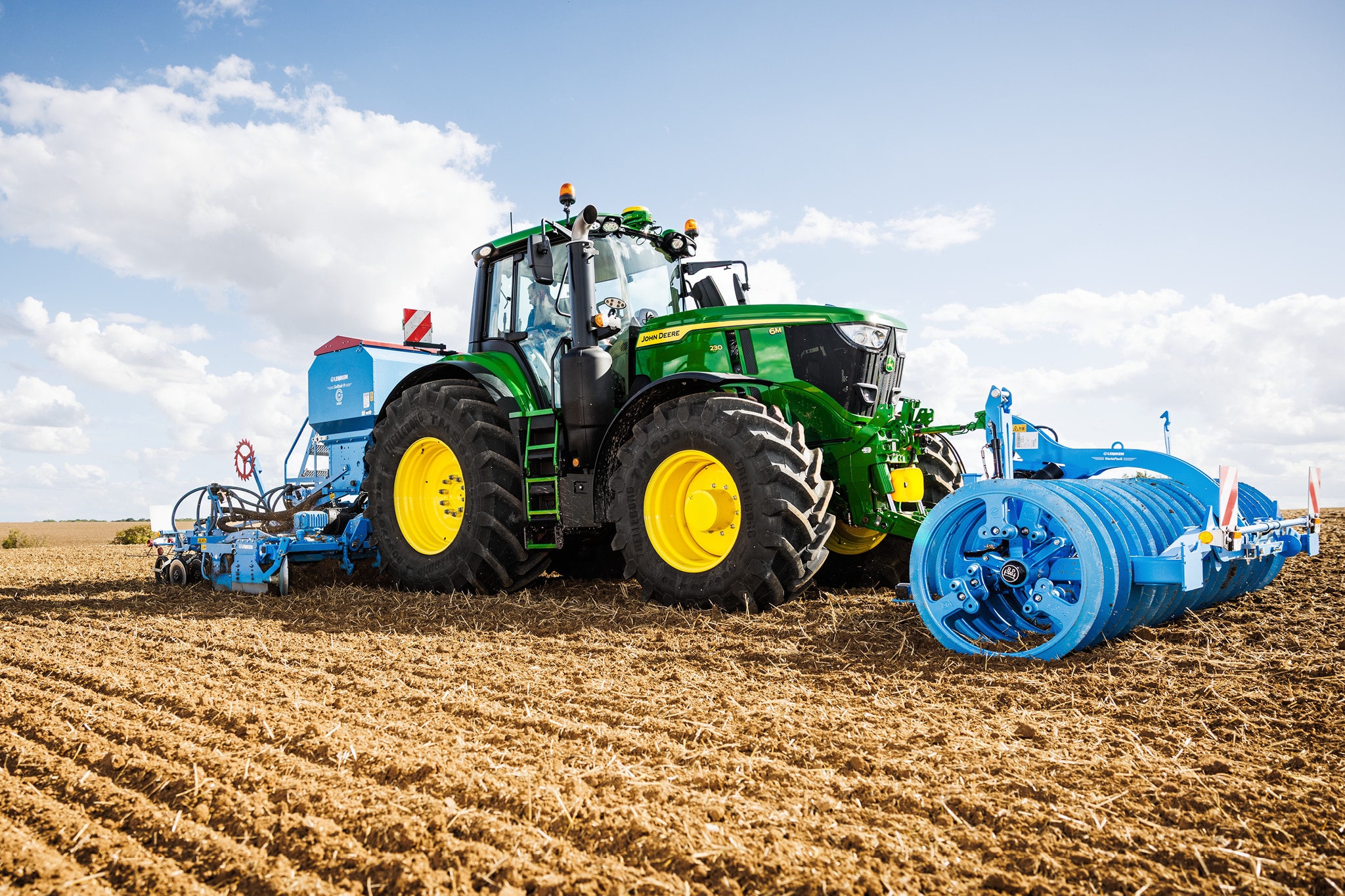 Traktor John Deere serije 6M obrađuje polje s priključenim velikim plavim valjkom.