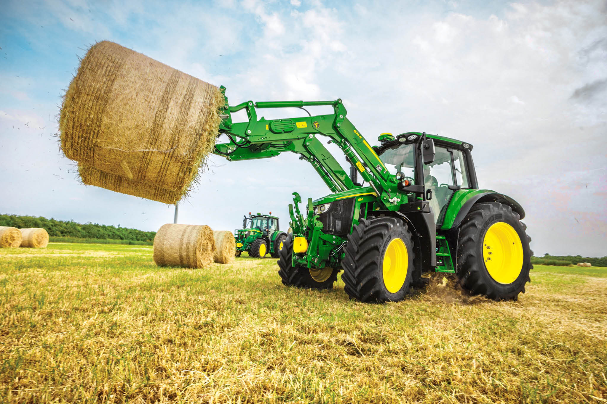 Traktor John Deere serije 6M podiže veliku okruglu balu sijena prednjim utovarivačem na otvorenom polju.