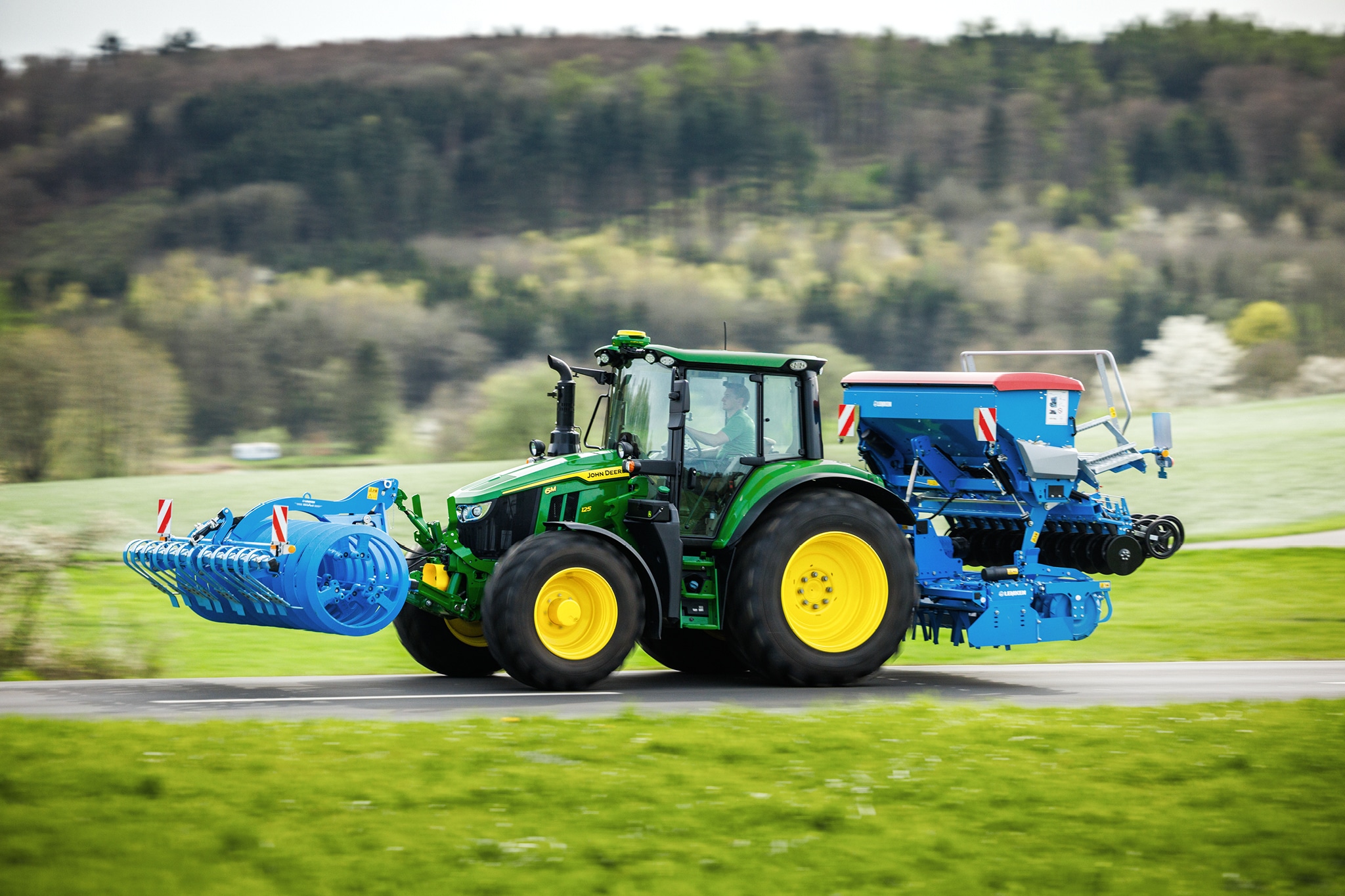 Traktor John Deere serije 6M koji prevozi sprijeda priključen alat i stražnju sijačicu duž ruralne ceste.