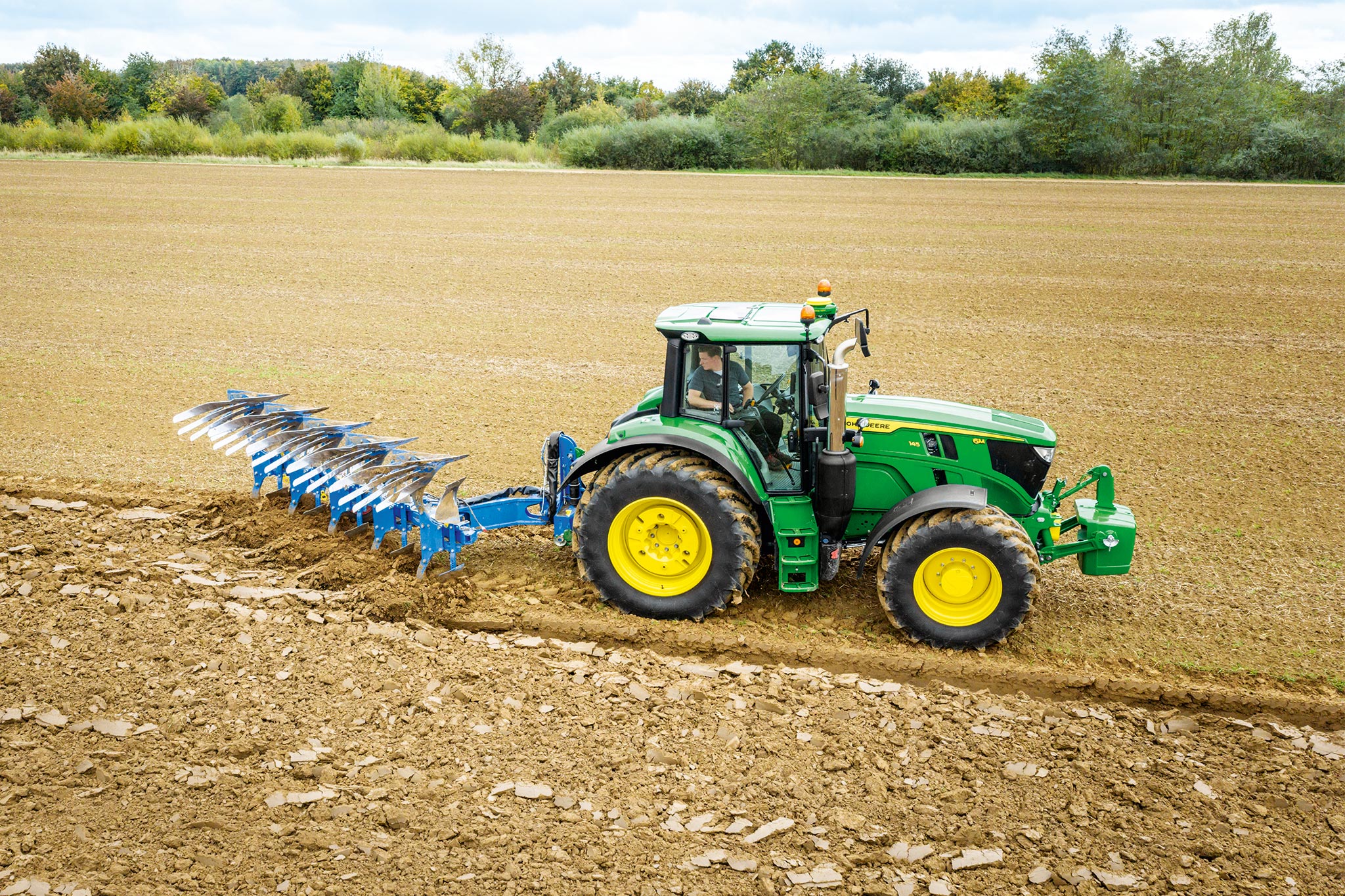 Traktor John Deere serije 6M radi na izoranom polju s alatom za prevrtanje priključenim na stražnjoj strani pod oblačnim nebom.