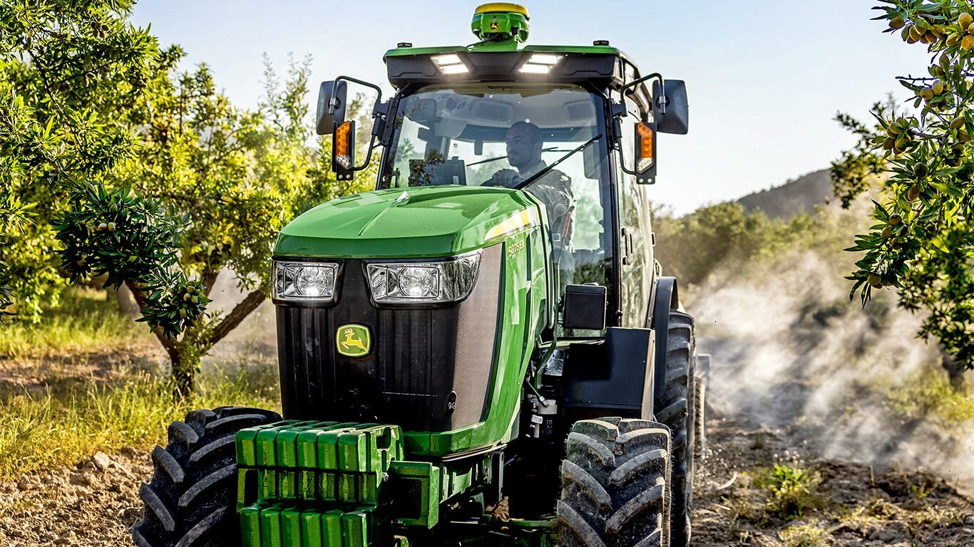 Zeleni i žuti John Deere 5075EN traktor vozi kroz voćnjak na prljavoj stazi, okružen voćkama i sunčevom svjetlošću.