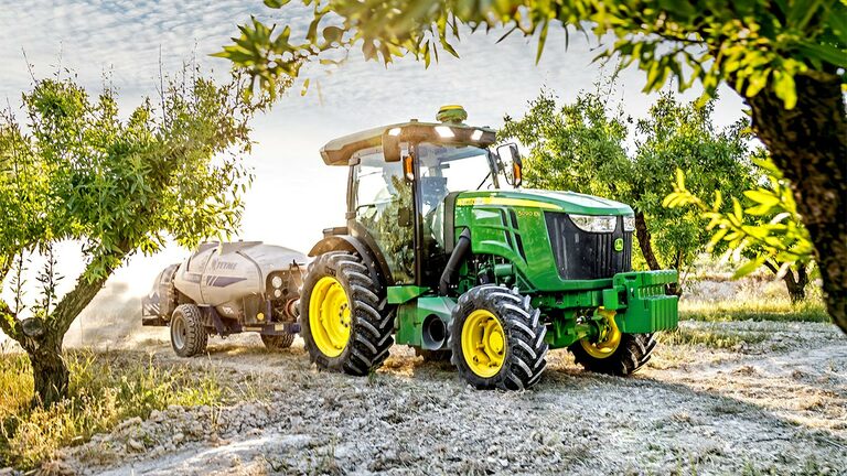 Zeleni i žuti John Deere traktor 5090EN vuče prskalicu kroz voćnjak na sunčan dan.