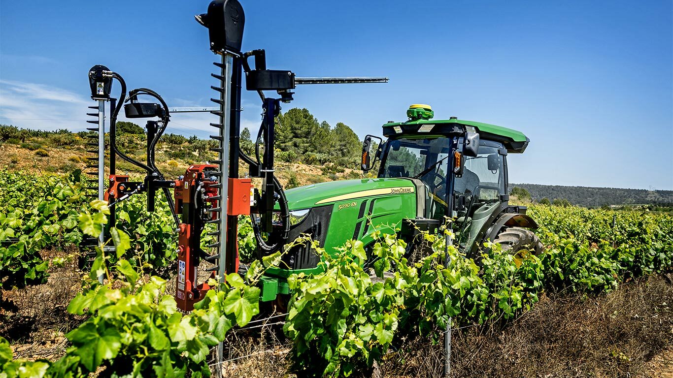 Zeleni i žuti John Deere 5090EN traktor opremljen alatima za obrezivanje koji rade u vinogradu pod vedrim plavim nebom.