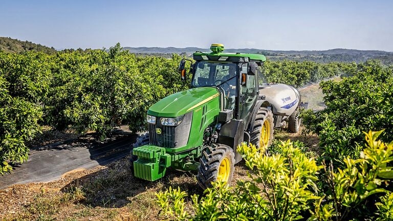 Zeleni i žuti John Deere 5105EN traktor vuče prskalicu u bujnom voćnjaku s brežuljkastom podlogom.