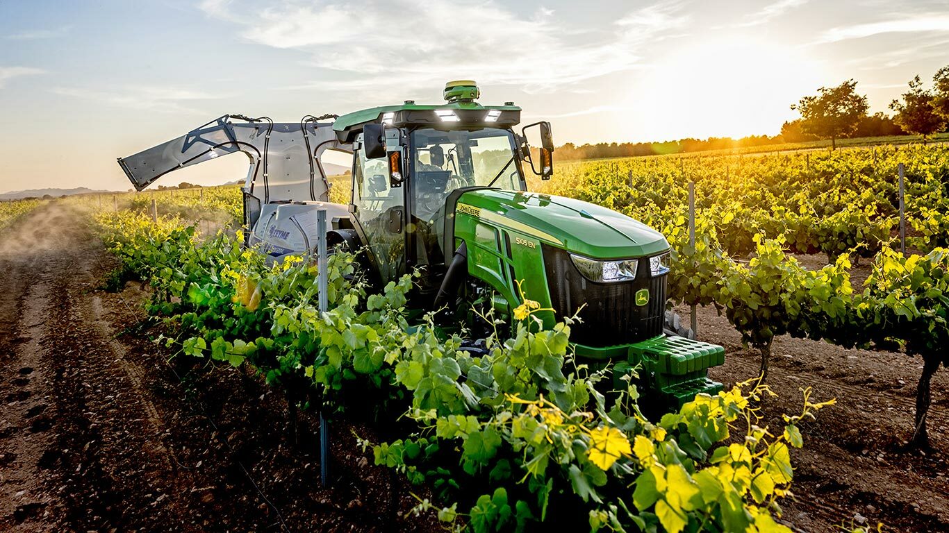 Zeleni i žuti John Deere 5105EN traktor radi u vinogradu u zalasku sunca, prskajući usjeve između redova vinove loze.