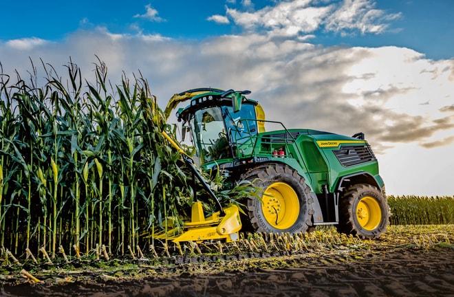 John Deere sakuplja kukuruz u polju pod plavim nebom s oblacima.