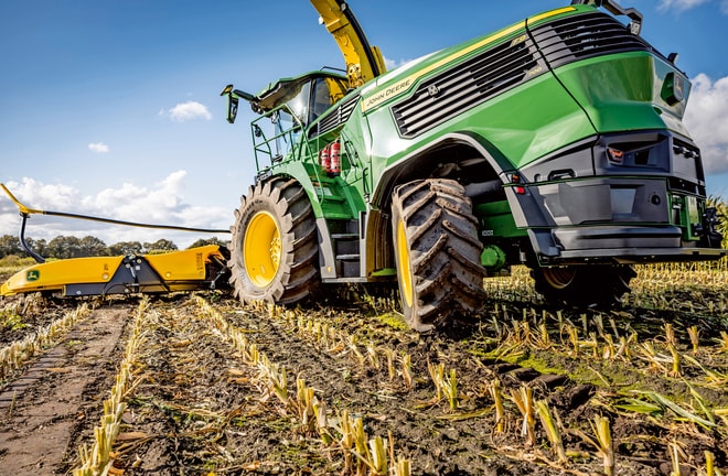 John Deere kombajn za stočnu hranu koji radi u žetvenom polju kukuruza, pokazujući velike gume stroja i mehanizam za rezanje na čistom plavom nebu.